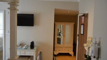 Een hotelkamer met een flatscreen televisie aan de muur, een houten kast met spiegel, een klein tafeltje met koffiezetapparaat, en een vaas met witte bloemen.
