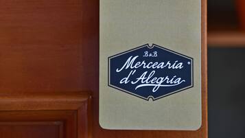 Uma porta com uma placa que diz 'POR FAVOR, ARRUME MEU QUARTO' e o logotipo da 'Merceria d'Alegria'.