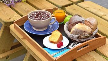 Una mesa de picnic de madera con una bandeja que contiene un tazón de cereal de chocolate, un vaso de jugo de naranja, una porción de pastel, dos frambuesas y una cesta de pan.