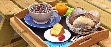 Une table de pique-nique en bois sur laquelle se trouve un plateau contenant un bol de céréales au chocolat, un verre de jus d’orange, une part de gâteau, deux framboises et un panier de pain.