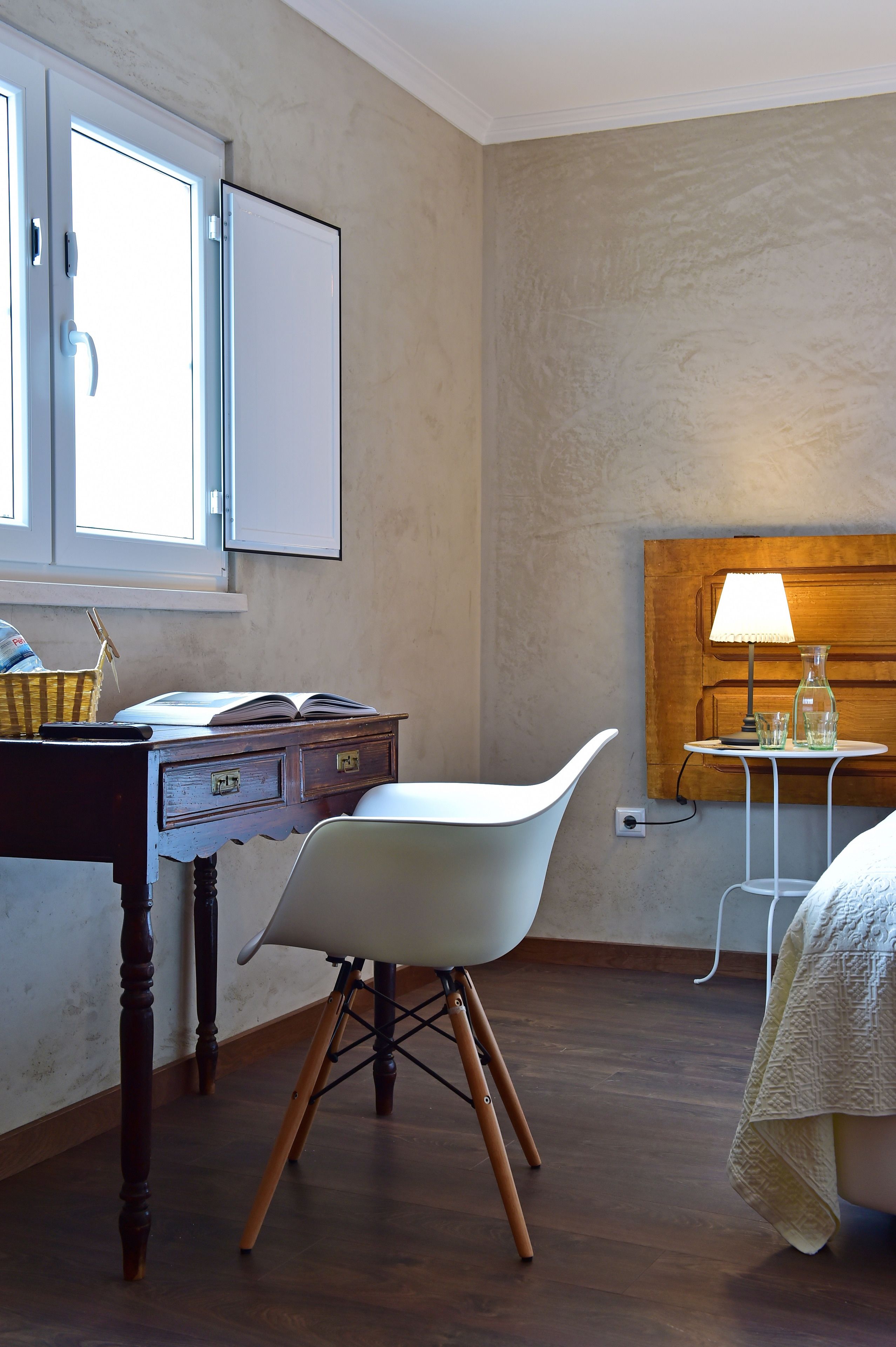 A room with a wooden desk, a white chair, a bed with a white cover, a lamp, and a wooden headboard.