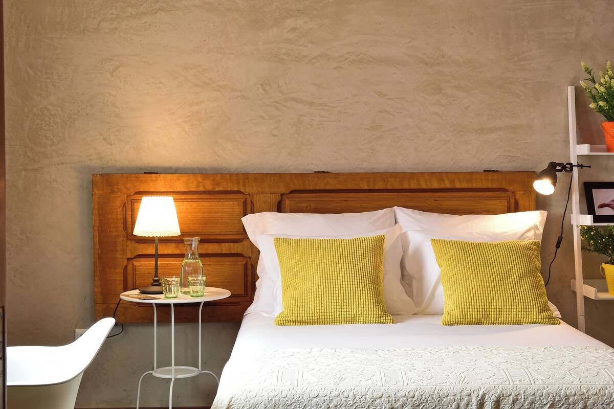 Une chambre à coucher avec une tête de lit en bois, un lit avec une literie blanche et jaune, une table de chevet avec une lampe, et une petite table d’appoint avec une lampe et une bouteille.
