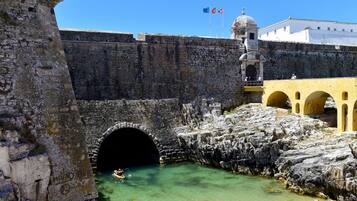 A historic stone bridge with a tunnel entrance, a yellow building with arches, and flags on a tower.