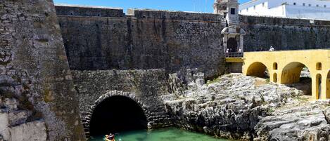 A historic stone bridge with a tunnel entrance, a yellow building with arches, and flags on a tower.