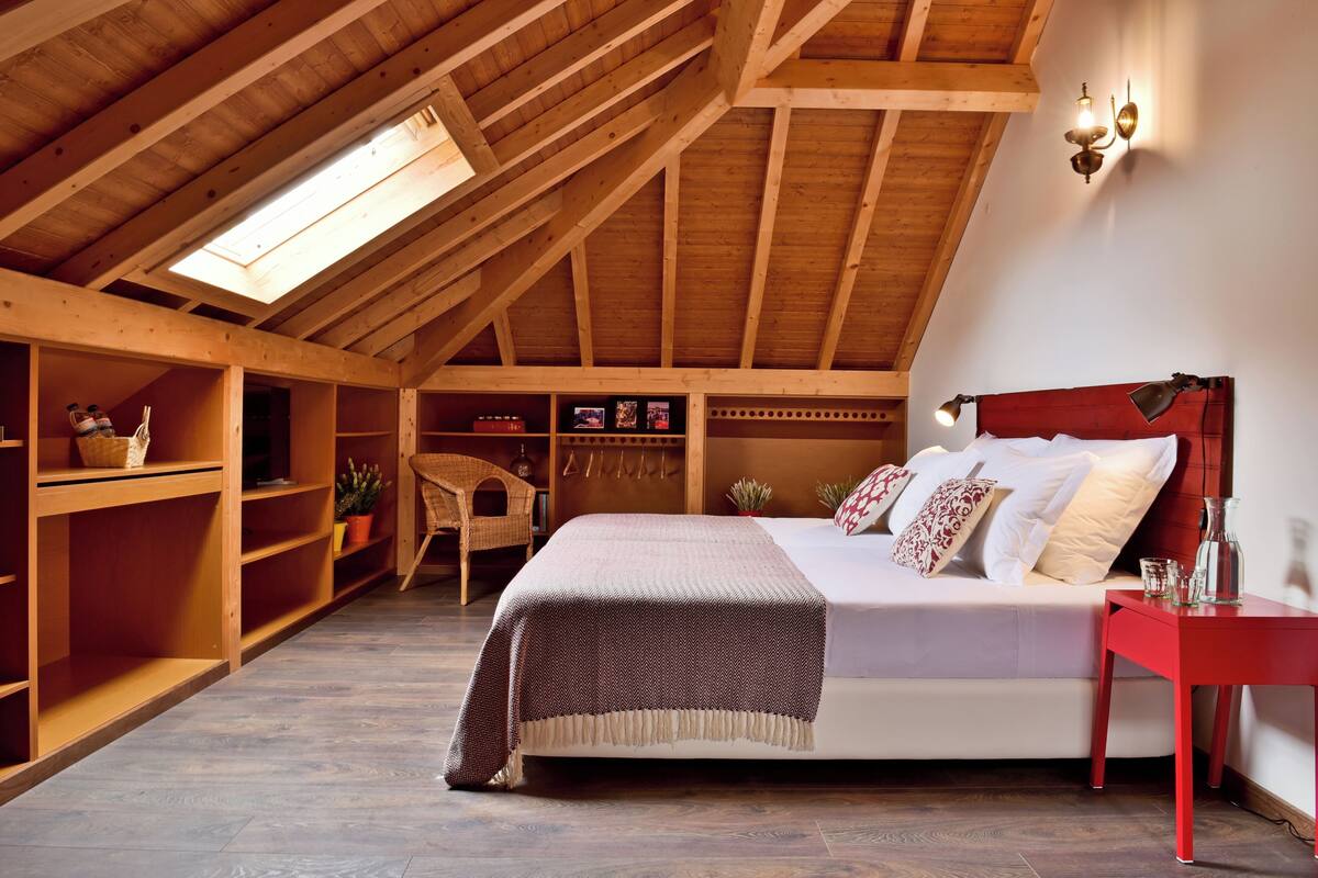 Une chambre avec un plafond en pente, des poutres en bois, un lit avec des oreillers, une table de chevet rouge et des étagères remplies de livres et de plantes.
