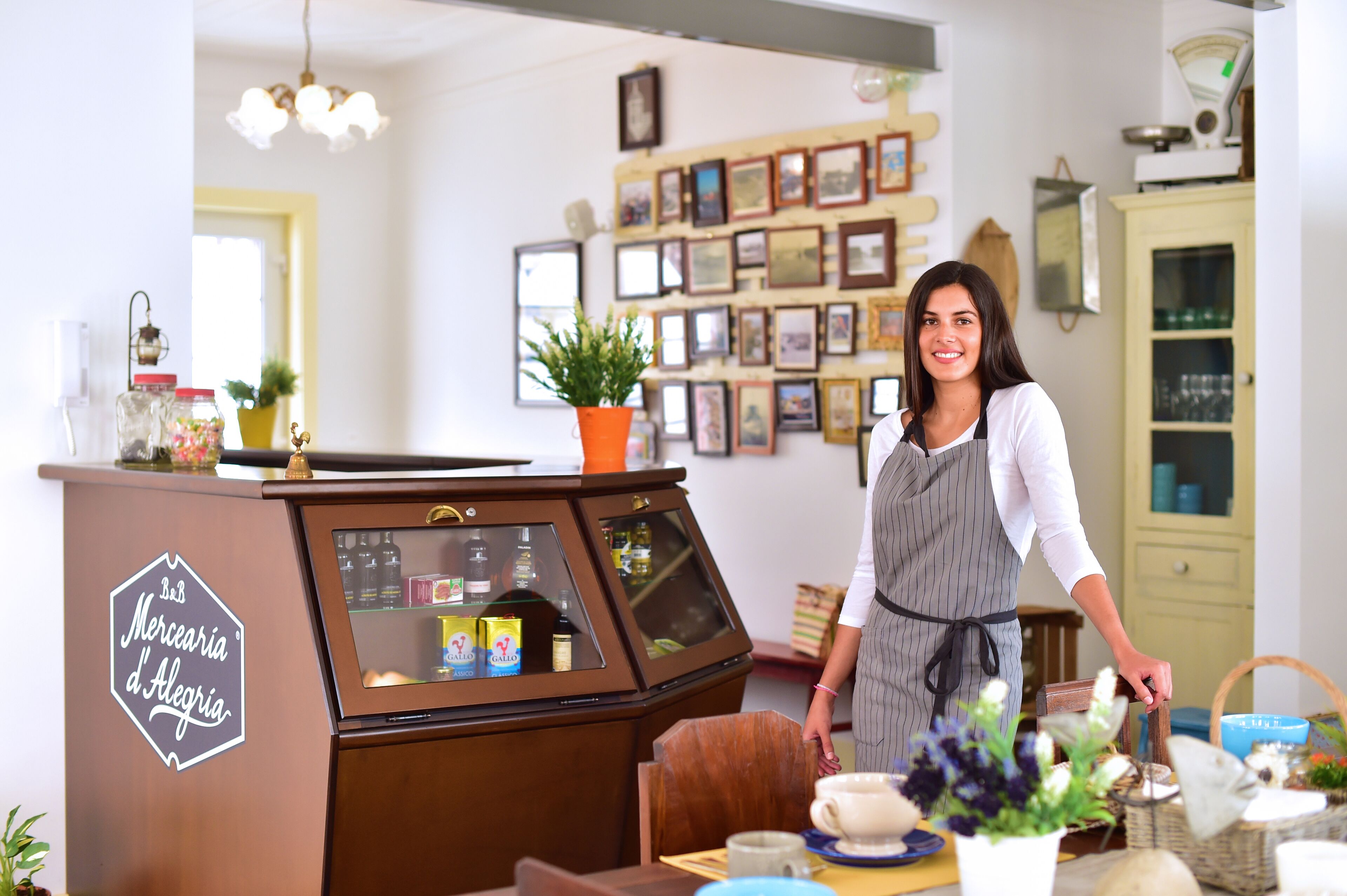 A vintage-style counter with a sign reading "Merceria d'Alegrias," a potted plant, and various items on display.