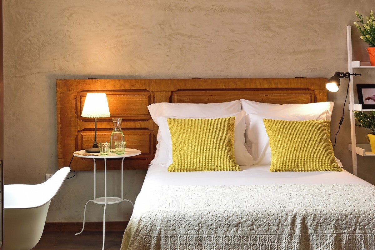 Un dormitorio con cabecera de madera, una cama con ropa de cama blanca y amarilla, una mesita de noche con lámpara, y una pequeña mesita auxiliar con lámpara y una botella.