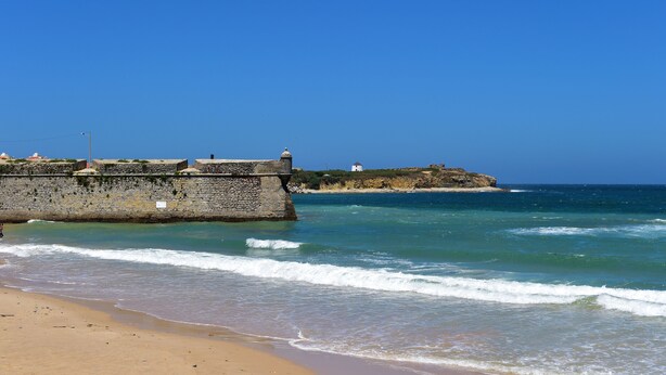 A coastal scene with a stone wall, beach, and rocky outcrop in the background. - Mercearia d'Alegria Boutique B&B (Peniche)