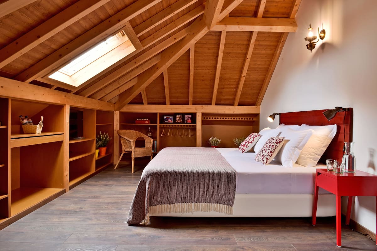 Une chambre avec un plafond en pente, des poutres en bois, un lit recouvert de draps blancs, une table de chevet rouge et un puits de lumière.
