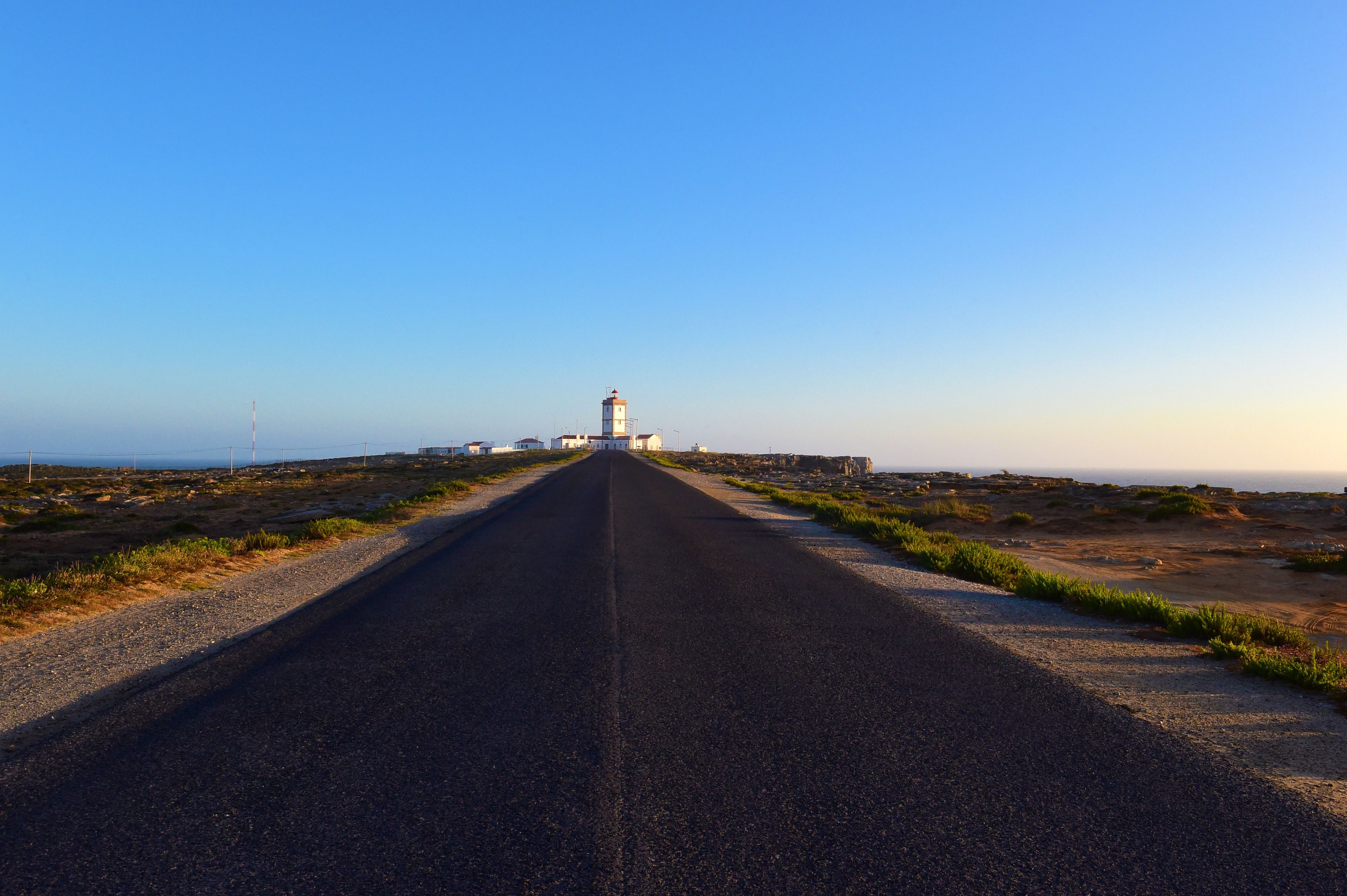 Een rechte weg die naar een vuurtoren leidt, onder een heldere lucht.