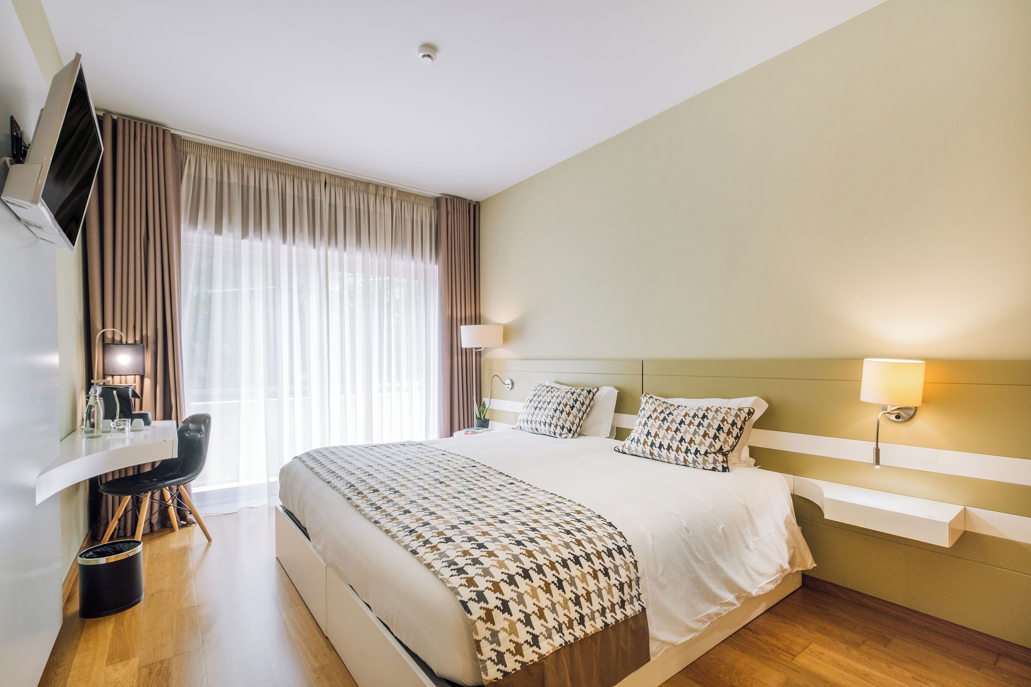 Ein Hotelzimmer mit einem großen Bett, Nachttischen, einem Schreibtisch mit Stuhl, einem an der Wand montierten Fernseher und einem Fenster mit Vorhängen.