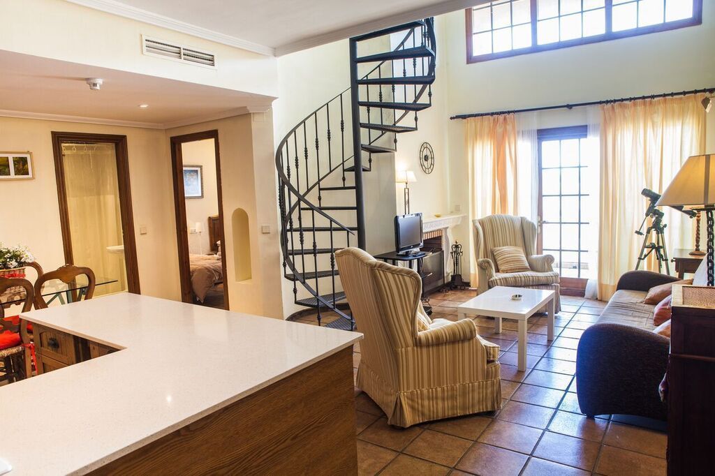 Un salón amplio con una escalera en espiral, un gran ventanal con cortinas y un comedor con mesa y sillas blancas.