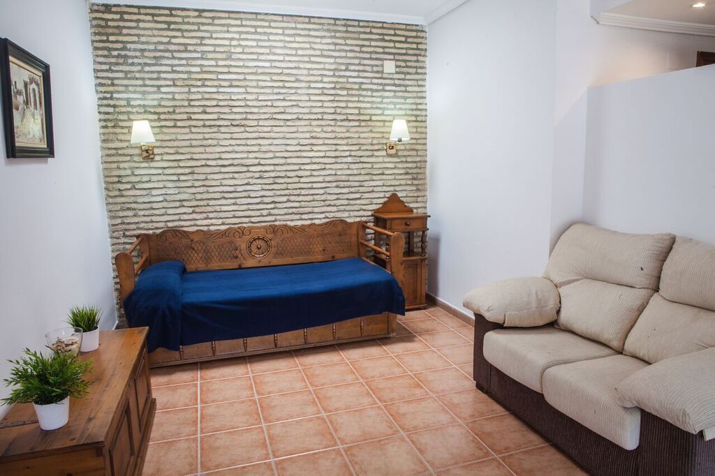 A room with a brick wall, a wooden sofa with a blue cover, a beige sofa, a wooden side table with plants, and two wall-mounted lamps.