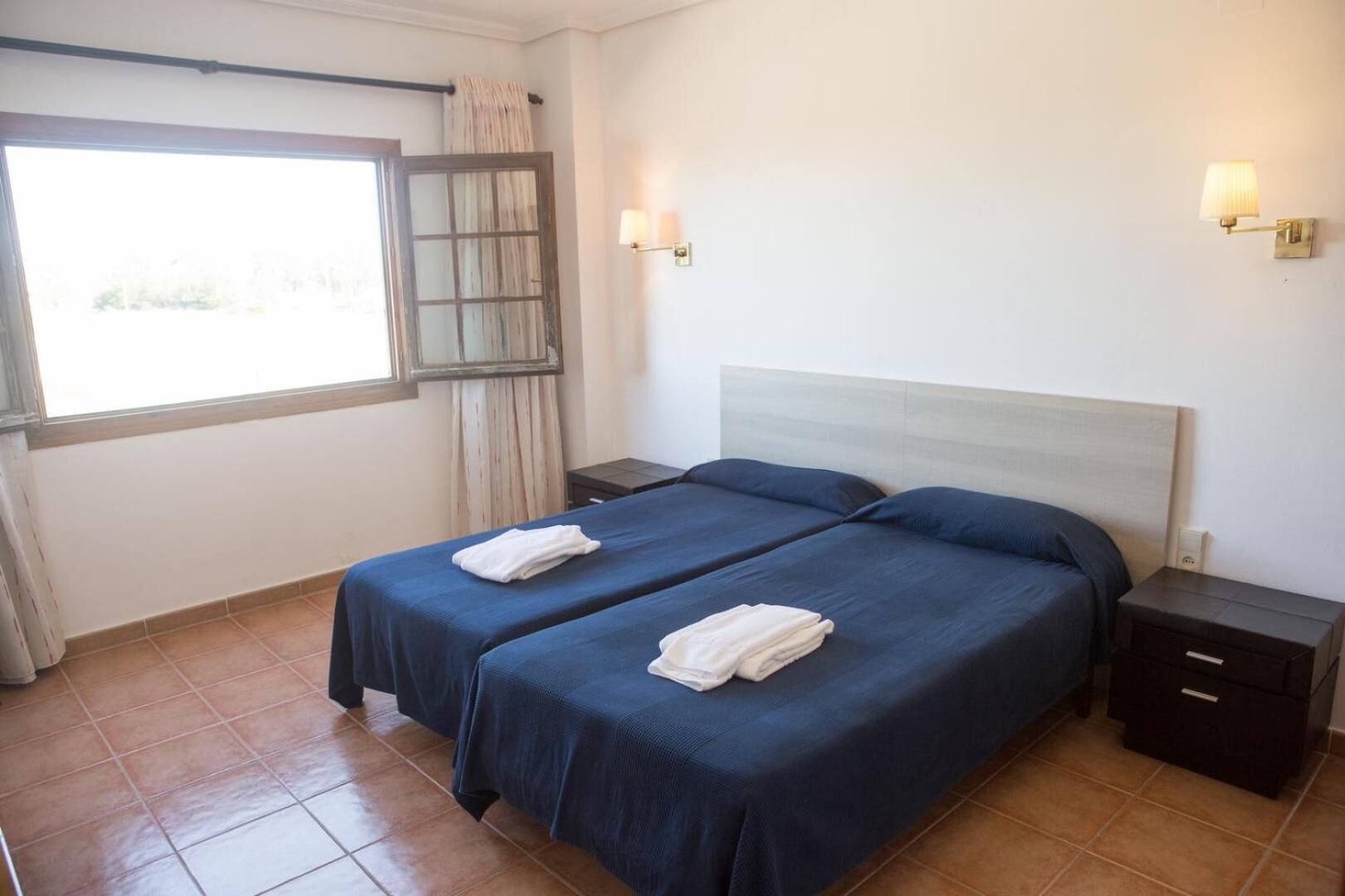 A hotel room with two beds, a window with curtains, a nightstand with a lamp, and a tiled floor.
