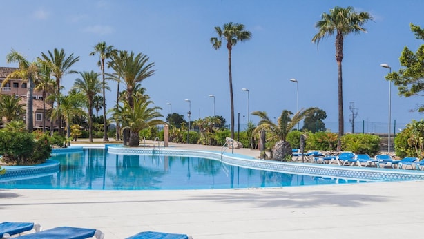 A swimming pool with blue water, surrounded by palm trees and blue lounge chairs. - Apartamentos Turísticos Bahía Sur (San Fernando)