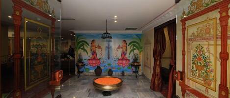 Un pasillo con una mesa redonda en el centro, flanqueado por decoraciones ornamentales en las paredes y un mural que muestra figuras con vestimenta tradicional.