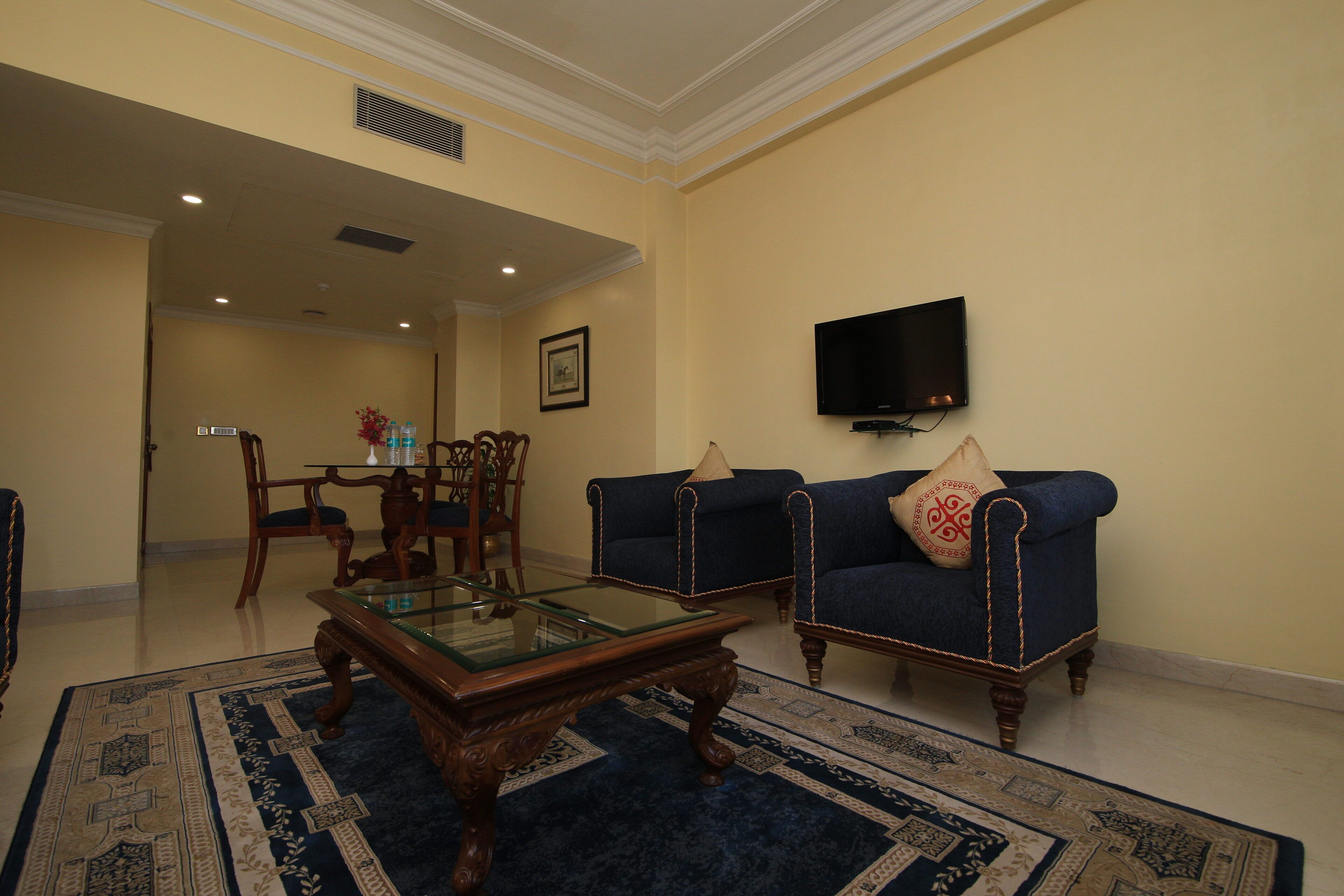 A living room with a glass coffee table, two blue sofas with decorative pillows, a flat-screen TV mounted on the wall, and a patterned rug.