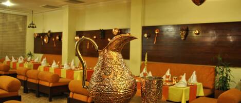 Un comedor con un gran jarro de cobre ornamentado y un vaso a juego sobre una mesa de madera, rodeado de asientos tapizados en naranja y mesas dispuestas con servilletas blancas y velas.