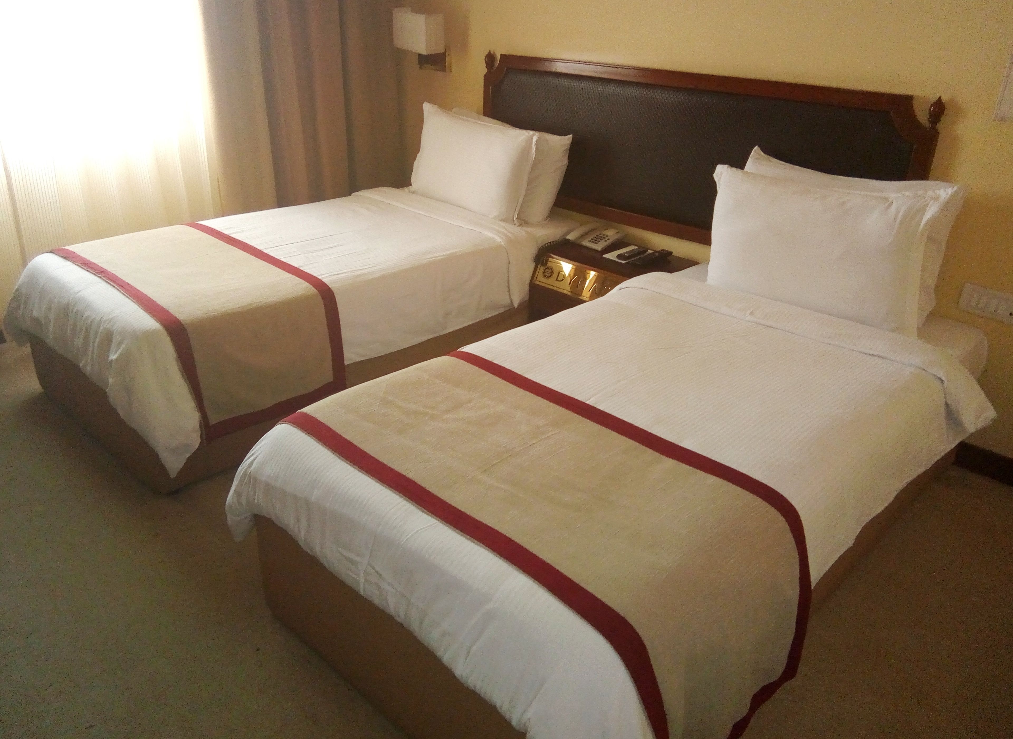 A hotel room with two single beds, each with a red-striped bedspread and white pillows. There is a wooden headboard, a bedside table with a lamp, and a telephone.