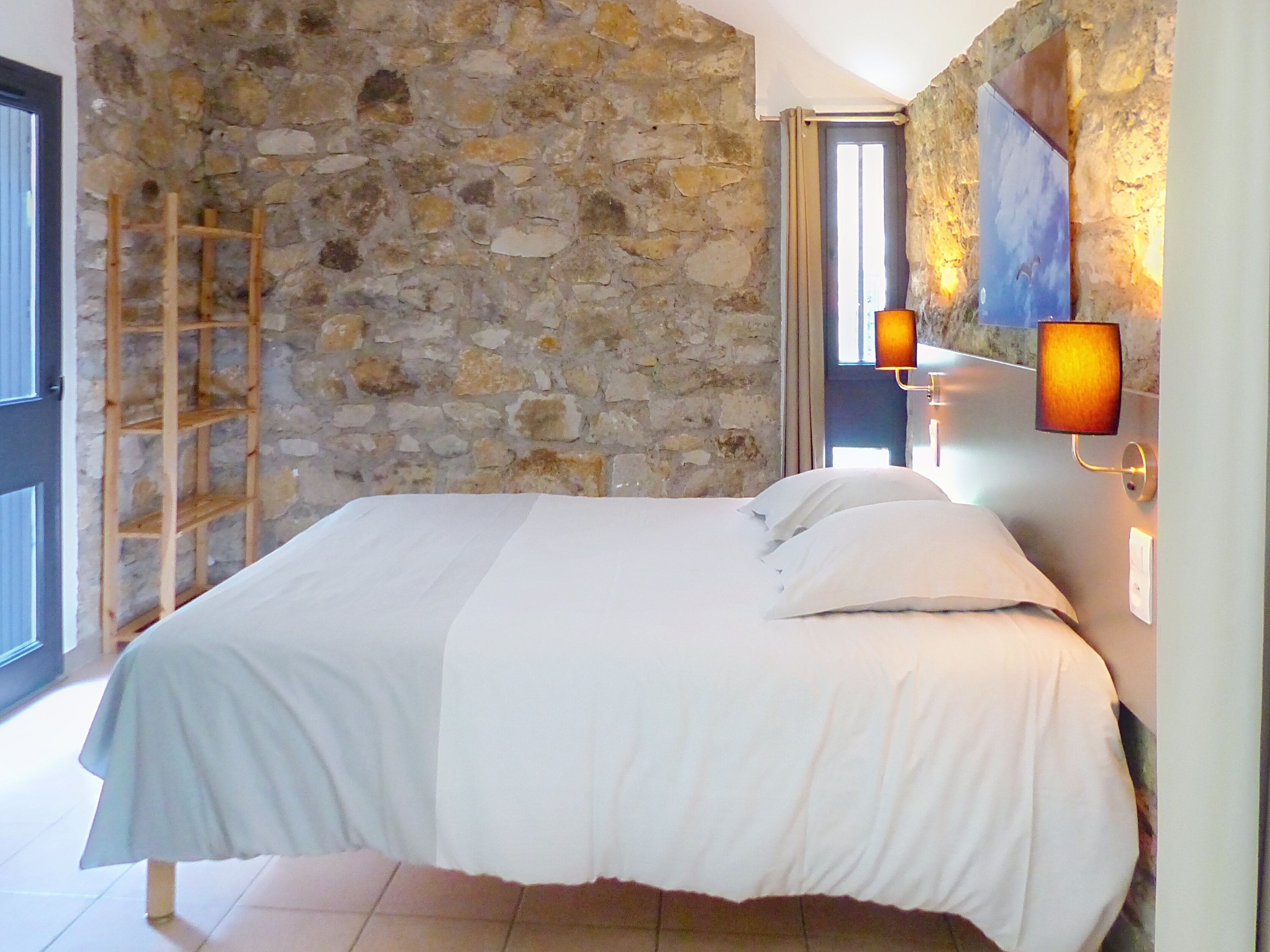 A bedroom with a stone wall, a wooden ladder, a bed with white bedding, and wall-mounted lamps.