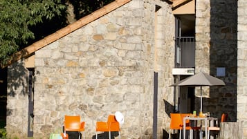 Un edificio de piedra con un balcón, mobiliario exterior con sillas anaranjadas y una mesa con sombrilla.