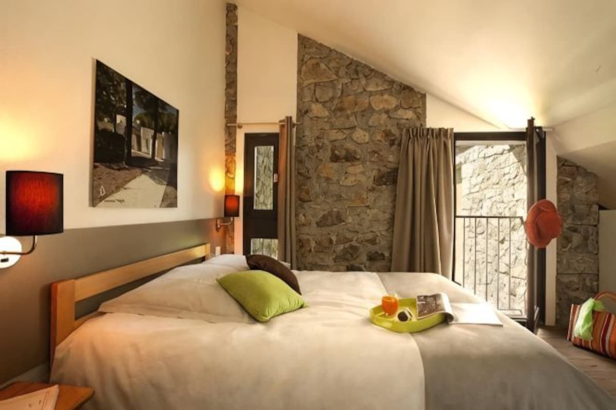 Un dormitorio con pared de piedra, una cama con almohadas, una mesita de noche, una lámpara y una ventana con cortinas.