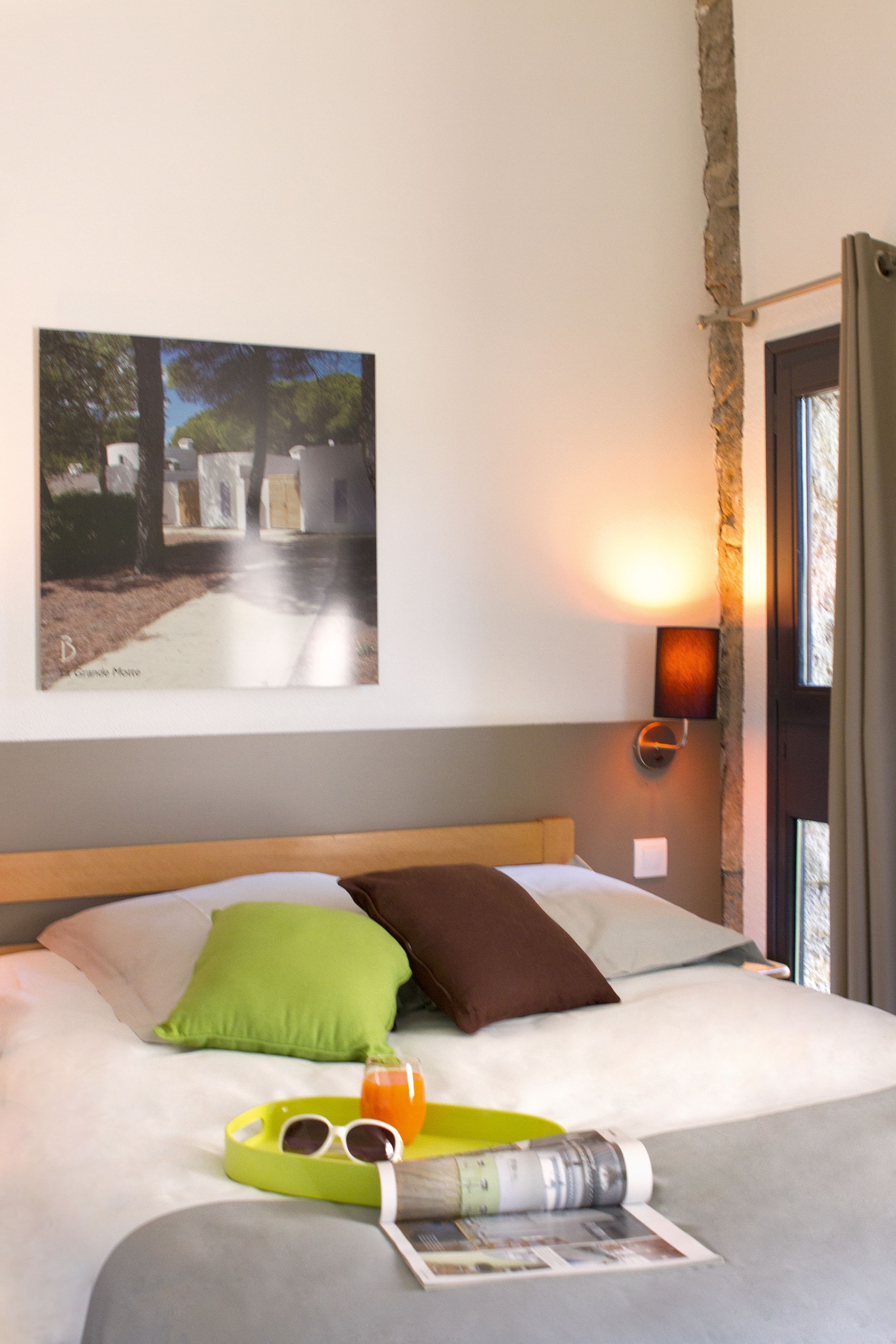 A hotel room with a bed, pillows, a tray with sunglasses and a drink, a wall-mounted picture, and a nightstand with a lamp.