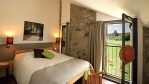 Un dormitorio con cama, pared de piedra, balcón con vistas y un sombrero colgado en la puerta.