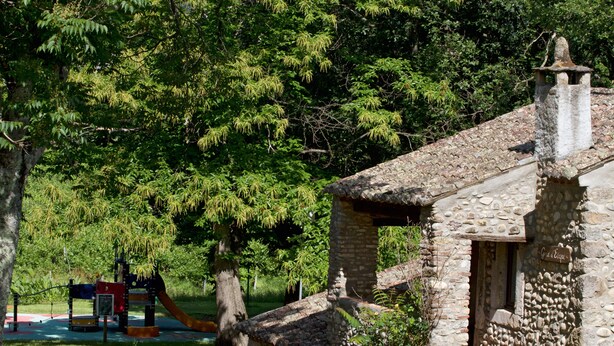Un edificio rústico de piedra con chimenea, rodeado de exuberante vegetación y un parque infantil.