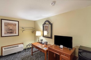 A hotel room with a desk, a television, a mirror, a framed picture, a lamp, and a heater. - Rodeway Inn Wauseon (Wauseon)
