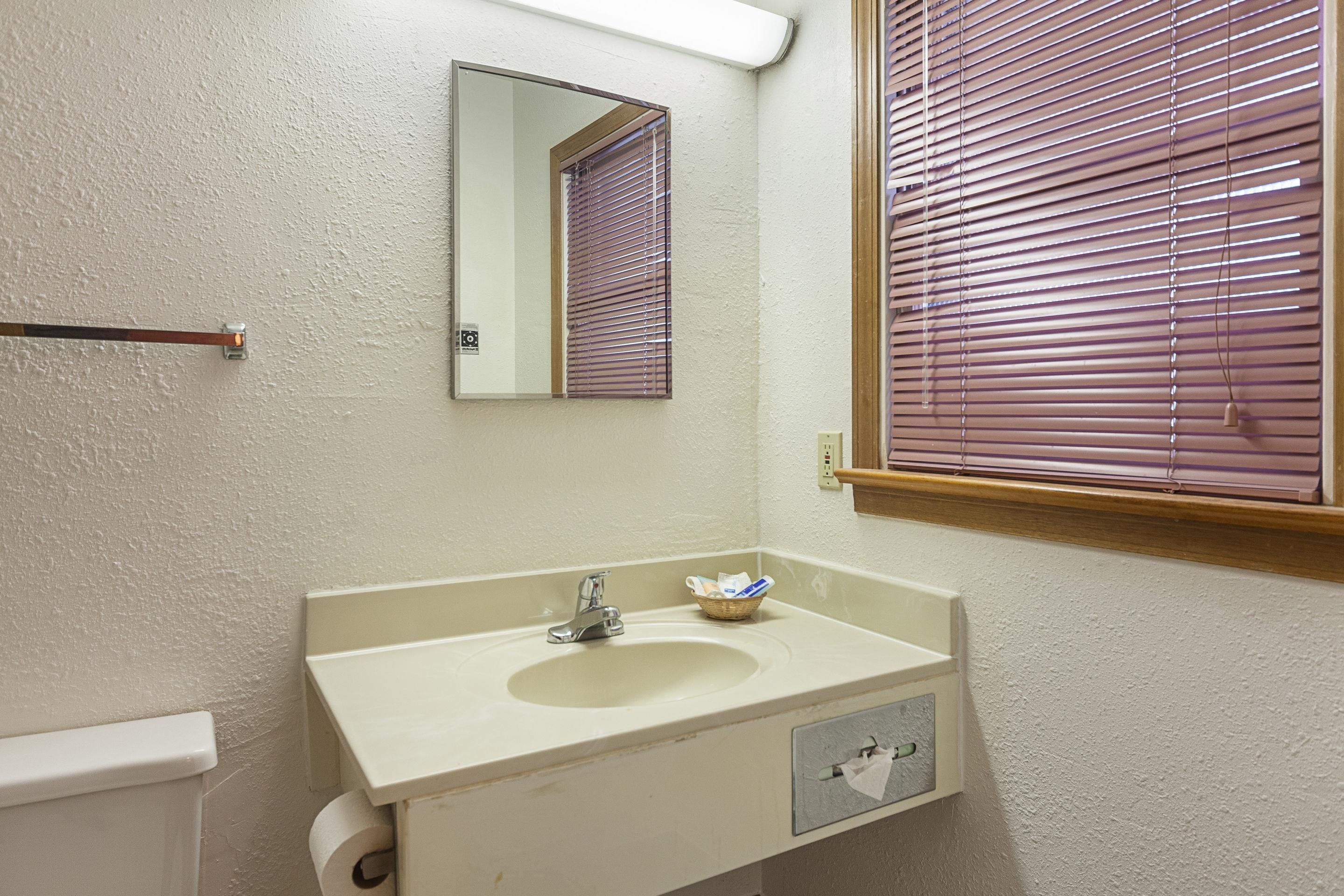 Un baño con lavamanos, espejo, ventana con persianas y dispensador de toallas.