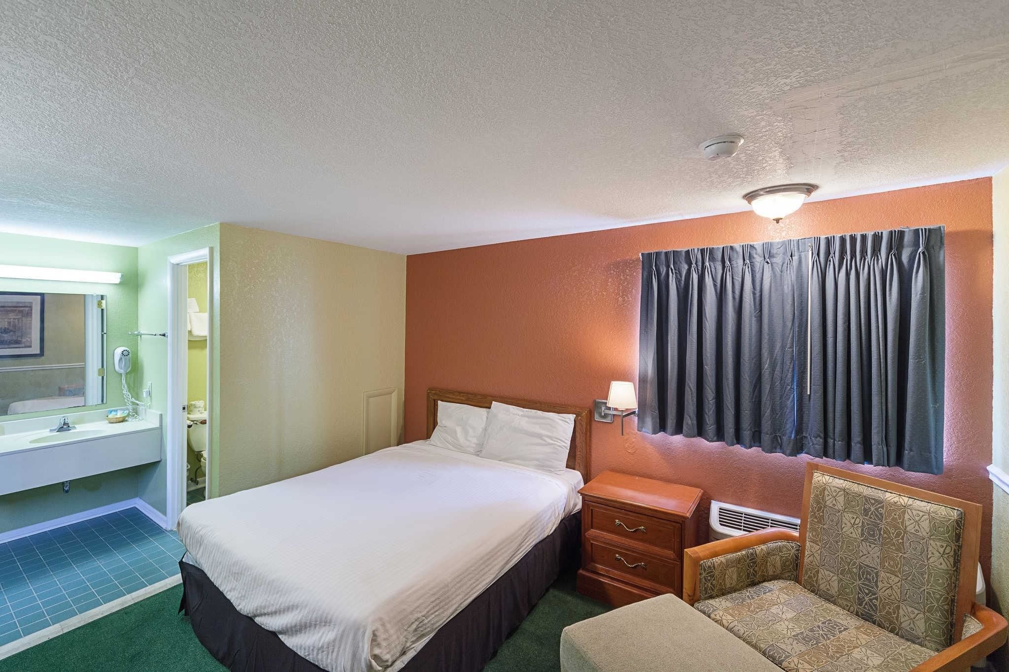 A hotel room with a bed, a nightstand, a chair, a bathroom with a sink and mirror, and a window with curtains.