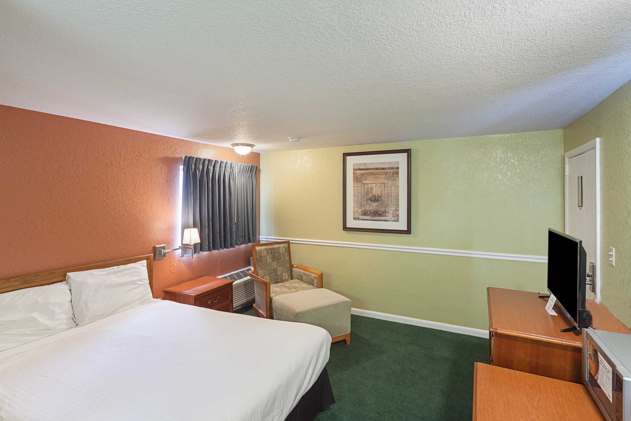 A hotel room with a large bed, a chair, a desk with a television, and a framed picture on the wall.