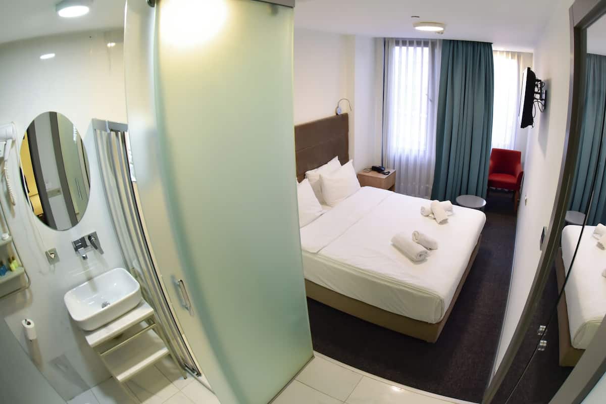 Habitación de hotel con cama, mesita de noche, televisor, una silla roja y baño con lavamanos y espejo.