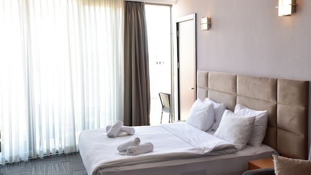 Quarto de hotel com uma cama grande, roupa de cama branca e bem arrumada, cabeceira e vista pela janela com cortinas finas.