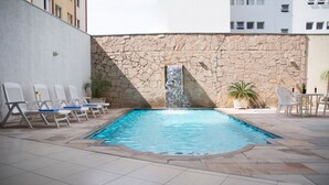 Una piscina exterior con pared de piedra y una cascada, rodeada de sillas de descanso y una mesa con sillas.