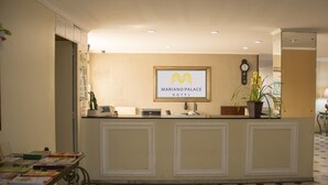 Una mesa de recepción con el logotipo enmarcado del Hotel Mariano Palace, un reloj y una planta en maceta.