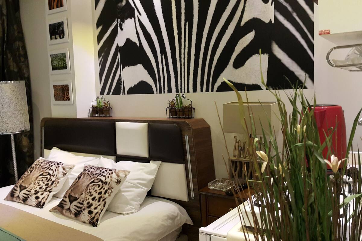 Une chambre avec un décor mural à rayures de zèbre, un lit avec des coussins à motifs léopard, une table de chevet avec une lampe et une valise rouge.