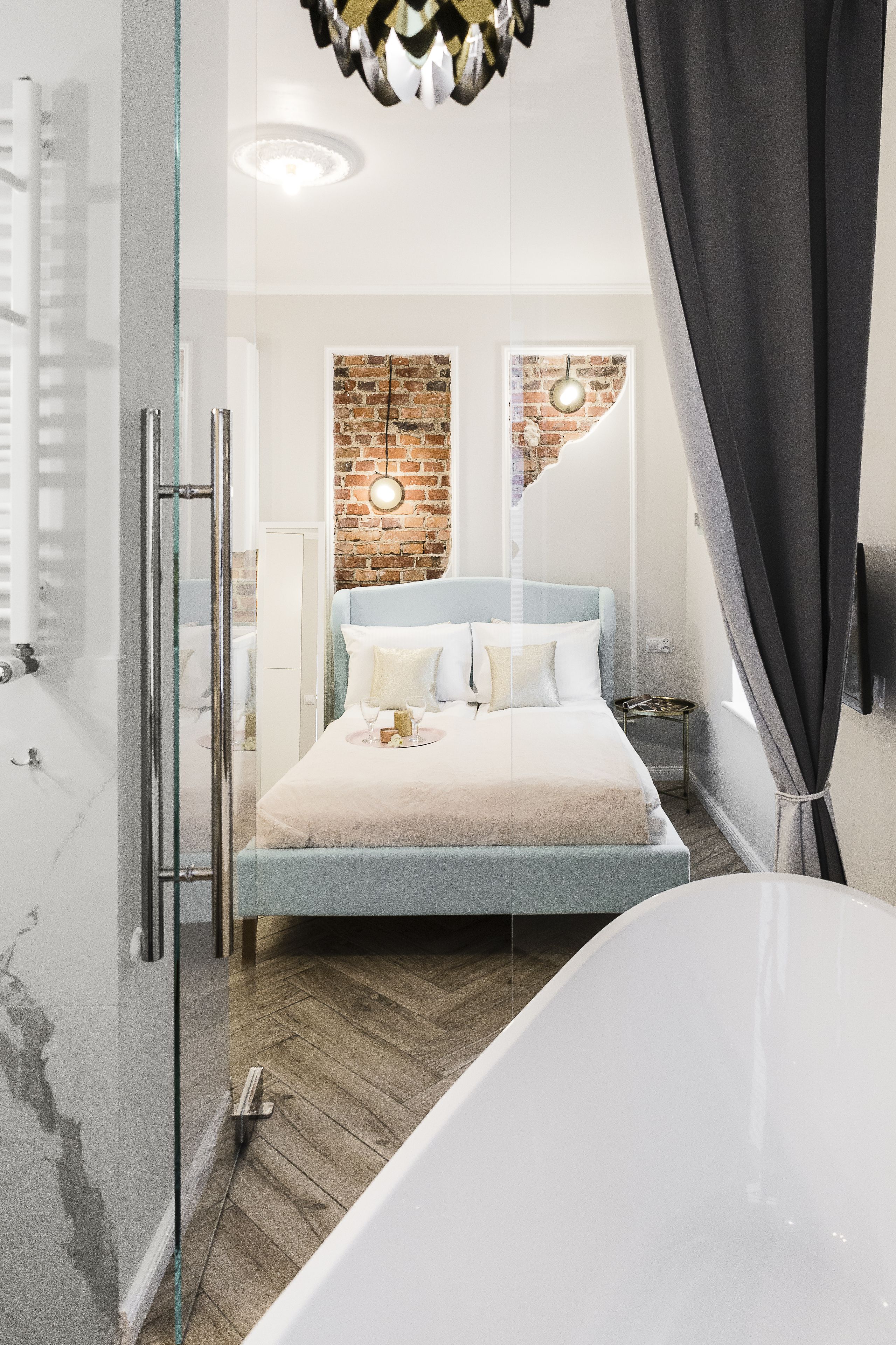 Une chambre moderne dotée d’une douche à parois de verre, d’un mur d’accent en briques, d’un grand lit avec une tête de lit bleu clair et d’une baignoire blanche.