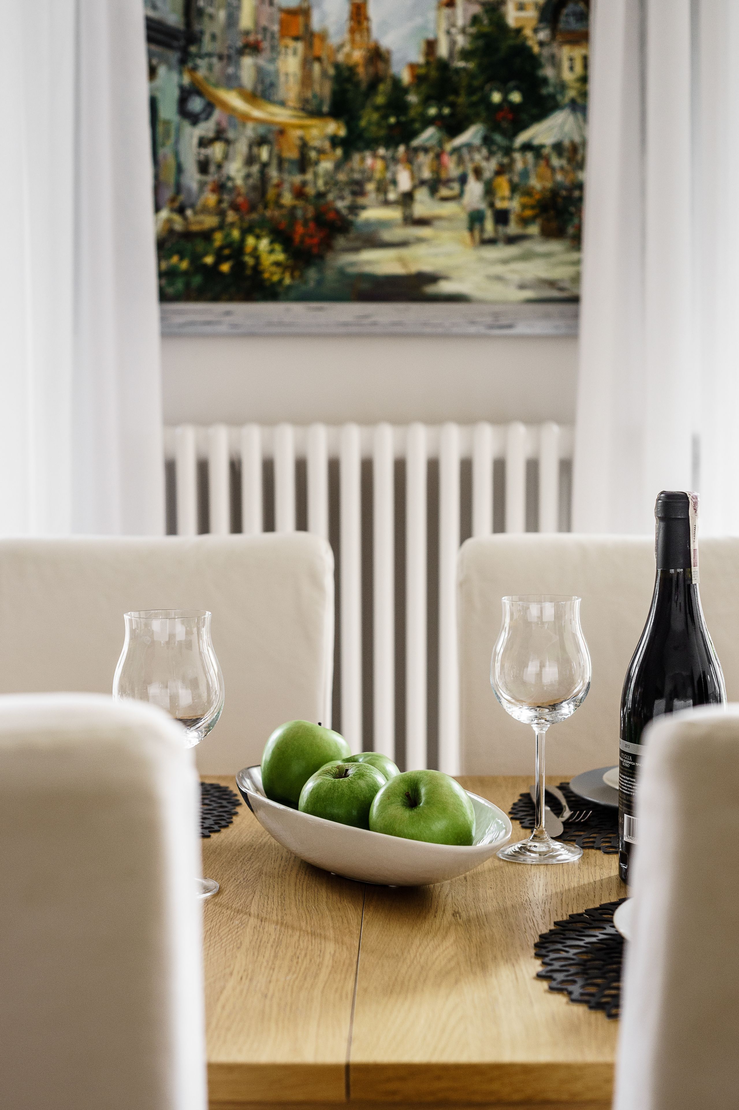 A dining table with a white bowl of green apples, two wine glasses, and a bottle of wine.