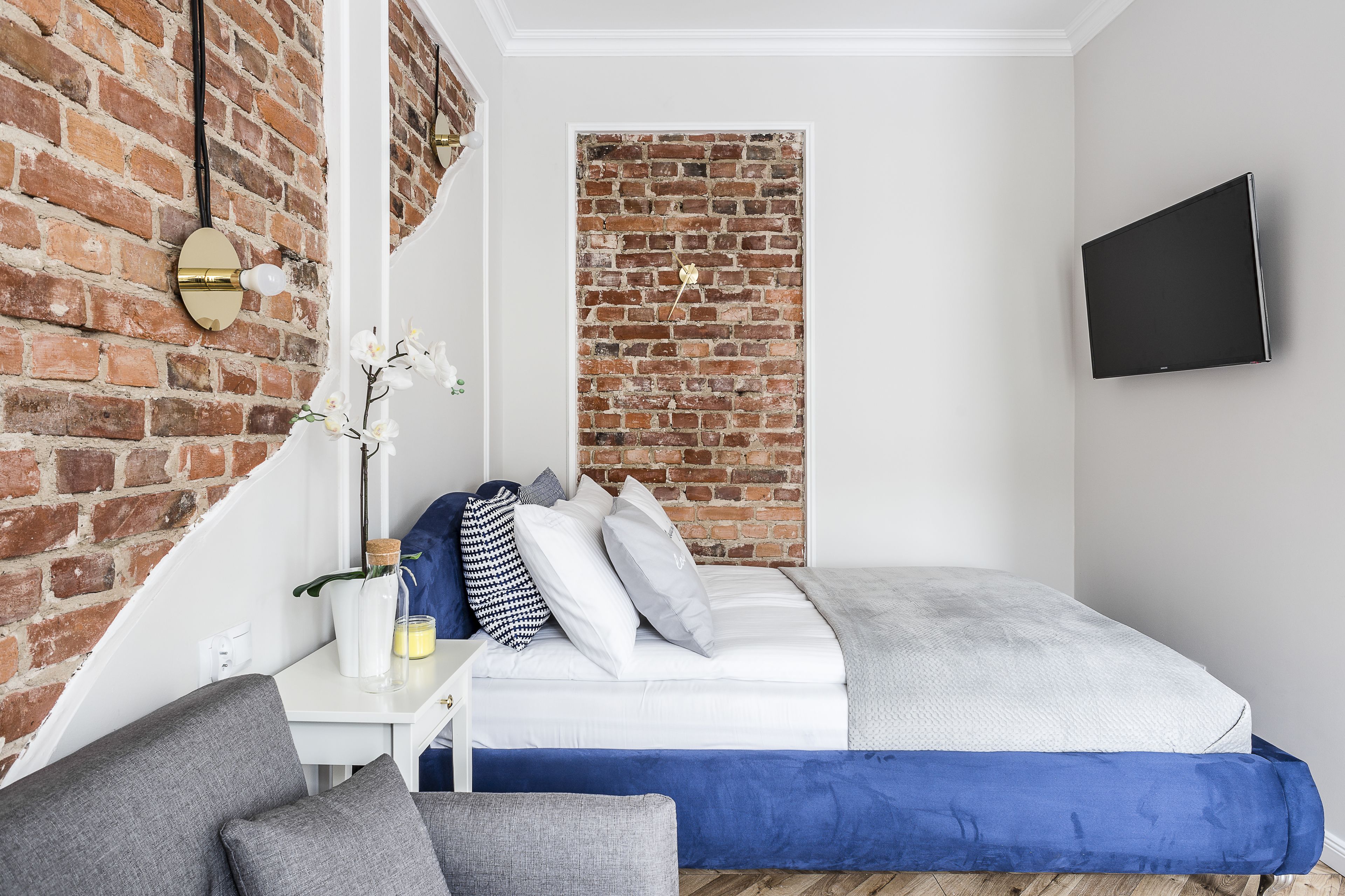 Un dormitorio moderno con pared de ladrillo, un televisor de pantalla plana, una cama blanca con ropa de cama azul y una mesita con una planta y una botella.