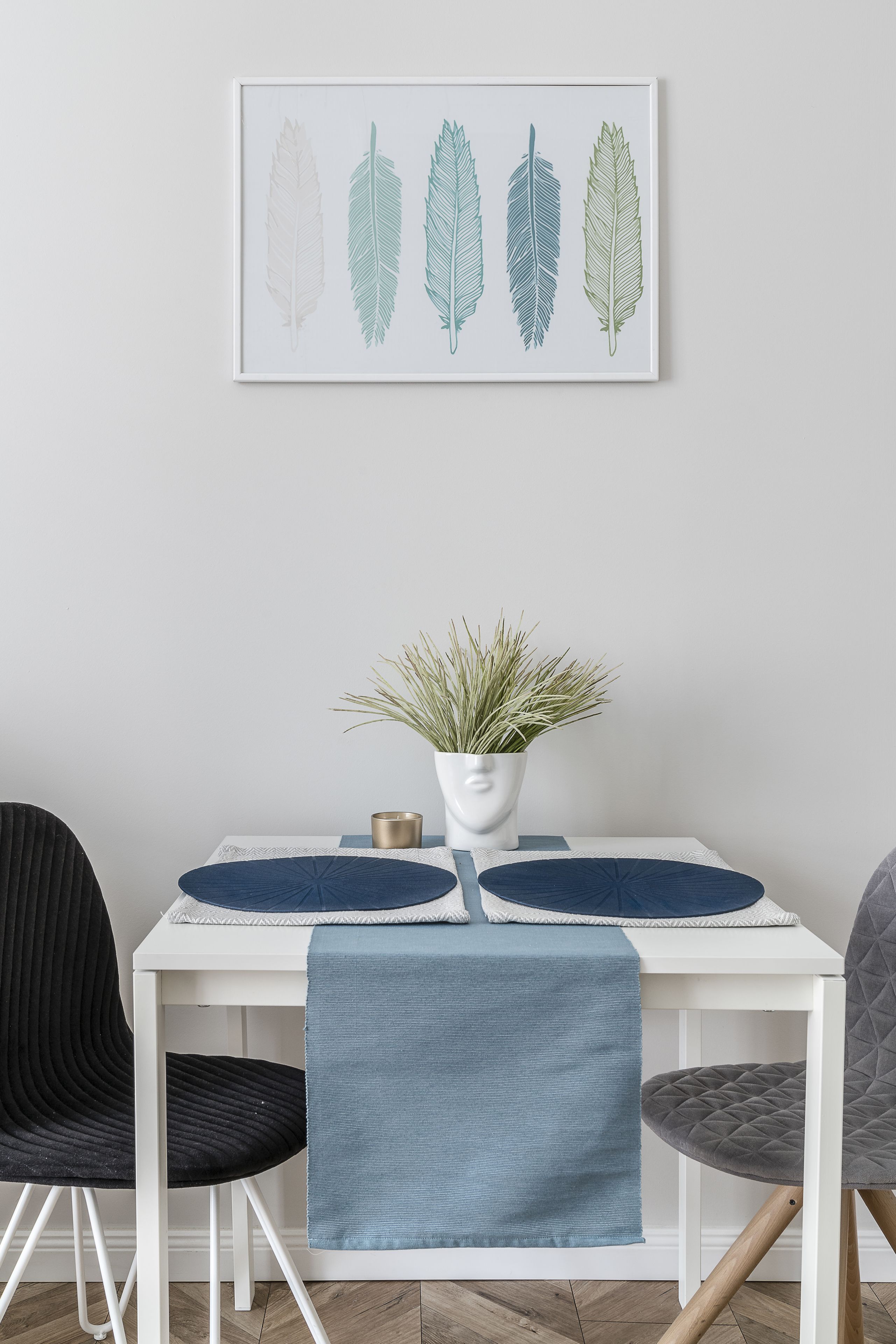 A dining area with a white table, two chairs, a blue table runner, a vase with a plant, and a framed artwork of leaves.