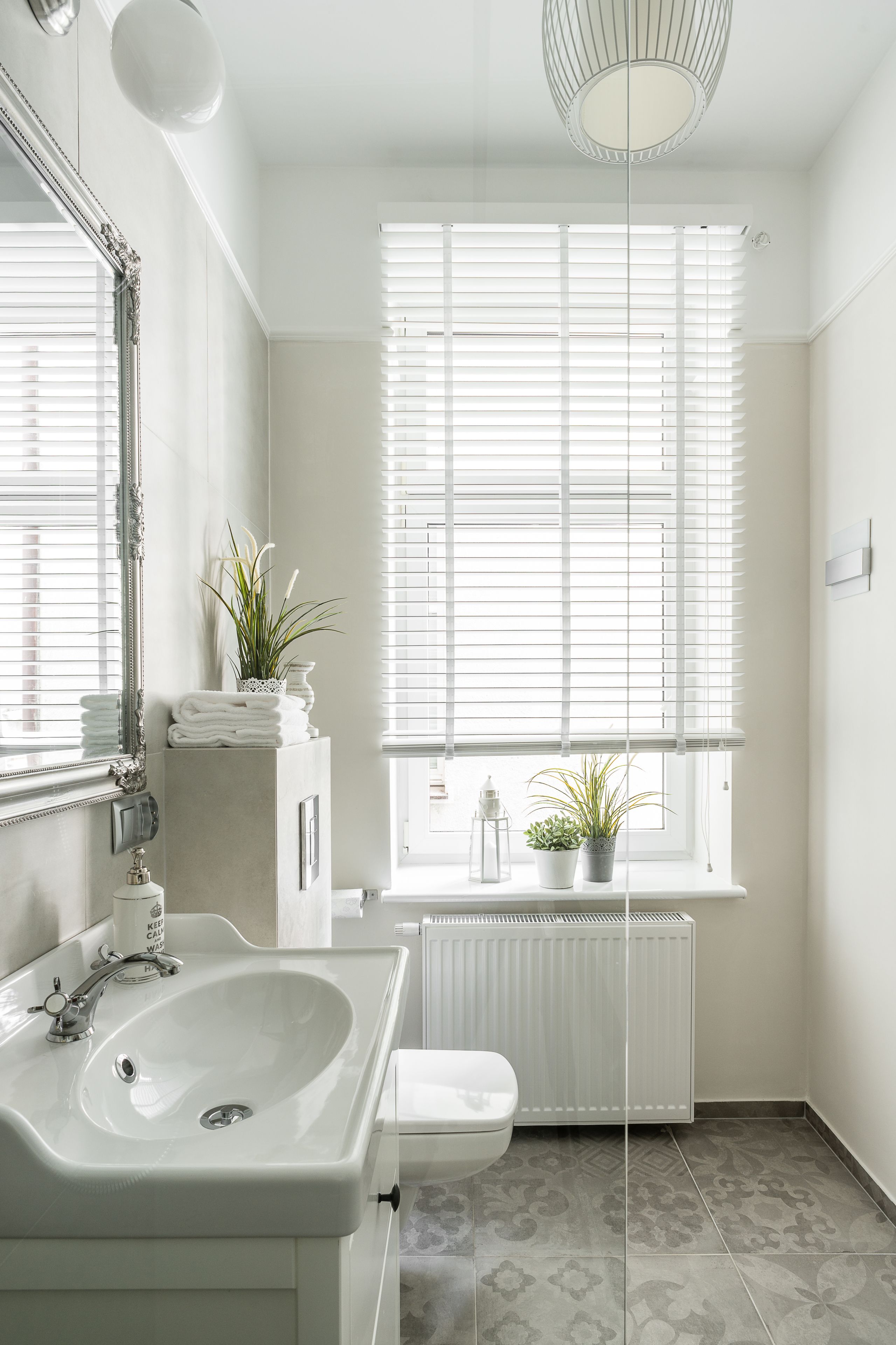 Un baño con lavamanos blanco, inodoro, radiador y ventana con persianas.