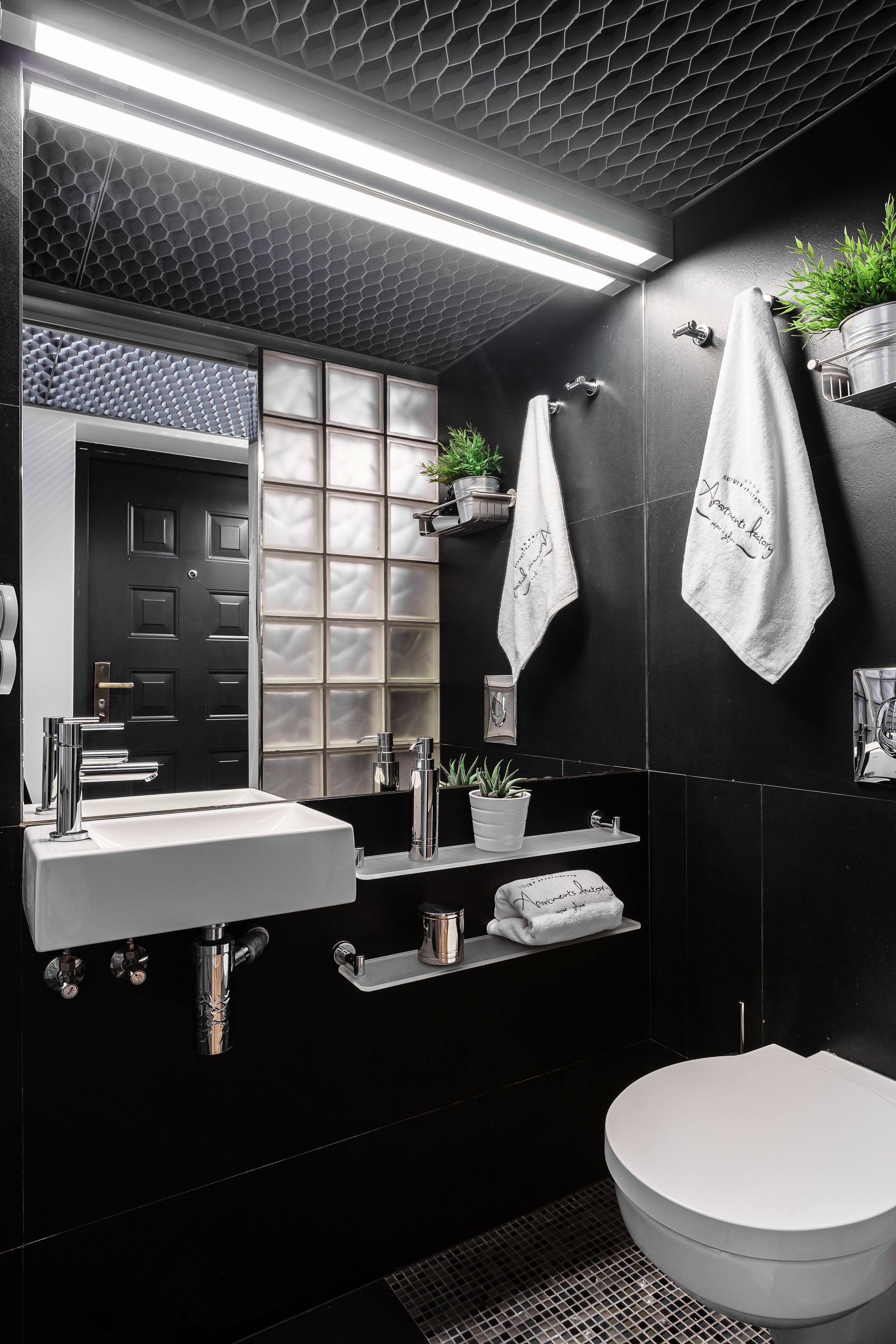 A modern bathroom with a black tiled wall, a white sink, a toilet, and a mirror. There are two towels hanging on hooks and a potted plant on the wall.