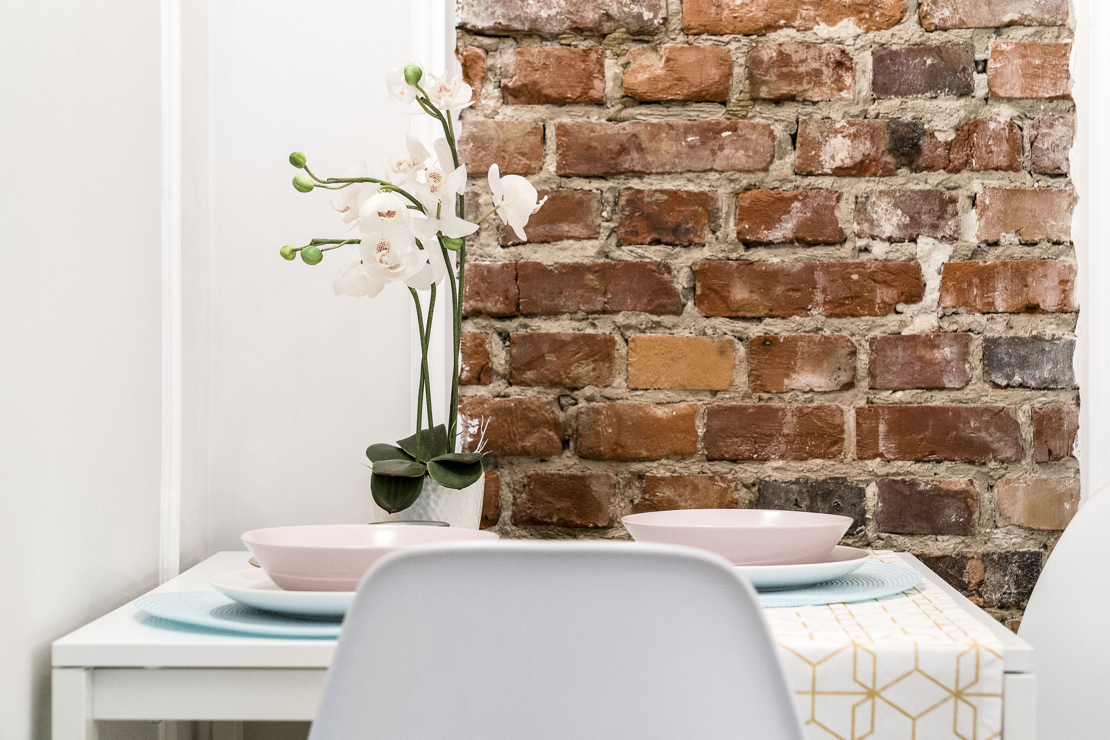 Un coin repas avec un mur en briques, une table blanche agrémentée de bols roses et un vase contenant des fleurs blanches.