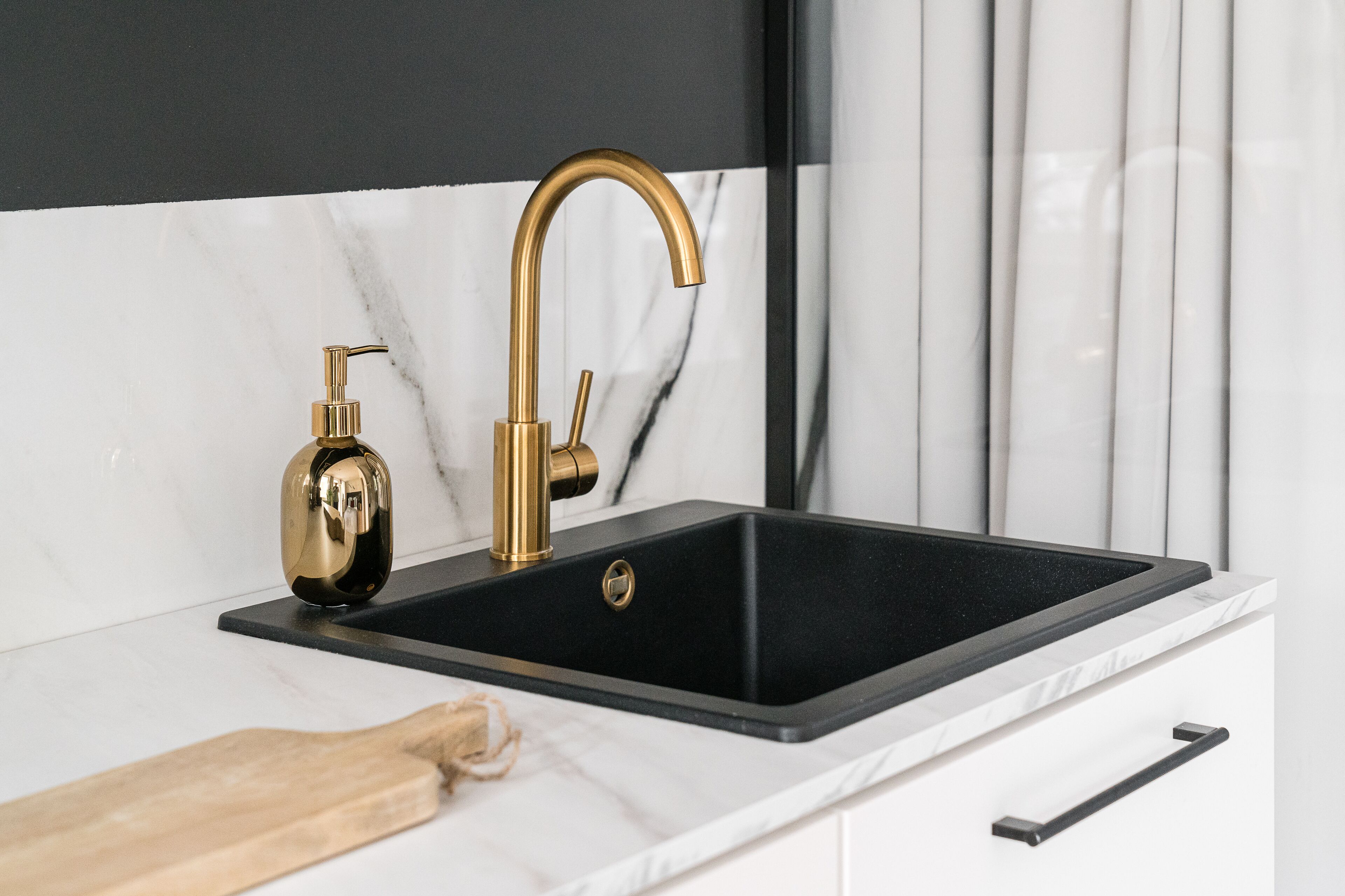 A kitchen with a black sink, gold faucet, and a wooden cutting board.