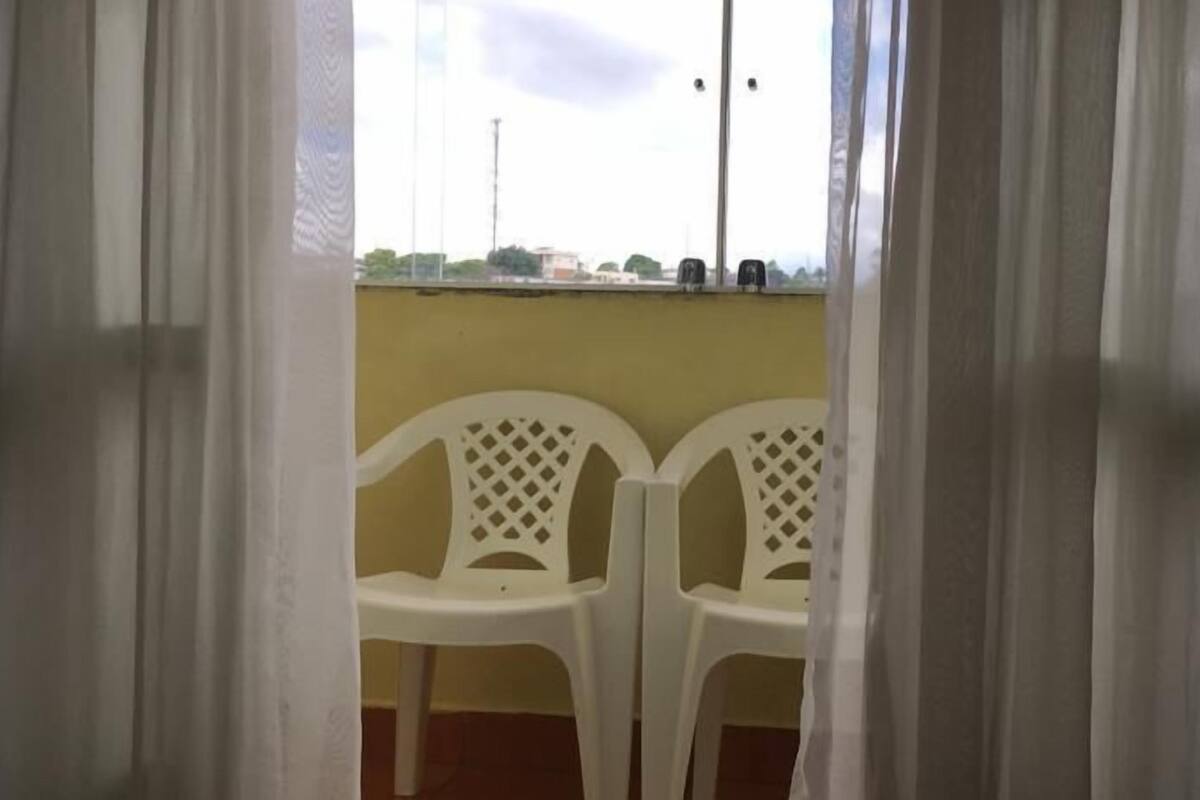 A view through a window with white curtains, revealing two white plastic chairs and a yellow wall.