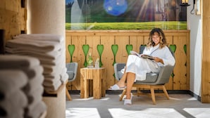 Una persona sentada en una silla leyendo un libro en una habitación con pared de madera y ventana con vista a un paisaje verde.