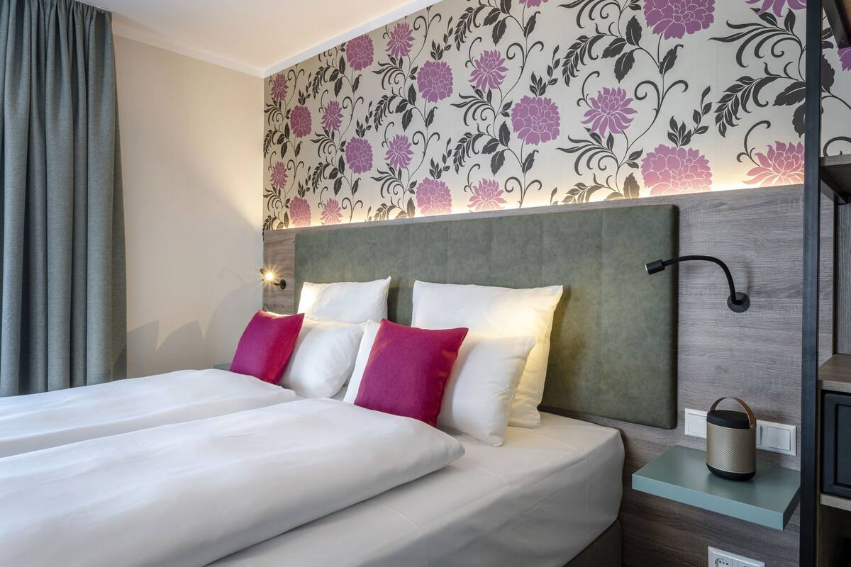 Habitación de hotel con cama, almohadas, mesita de noche, lámpara y un papel tapiz floral en la pared.