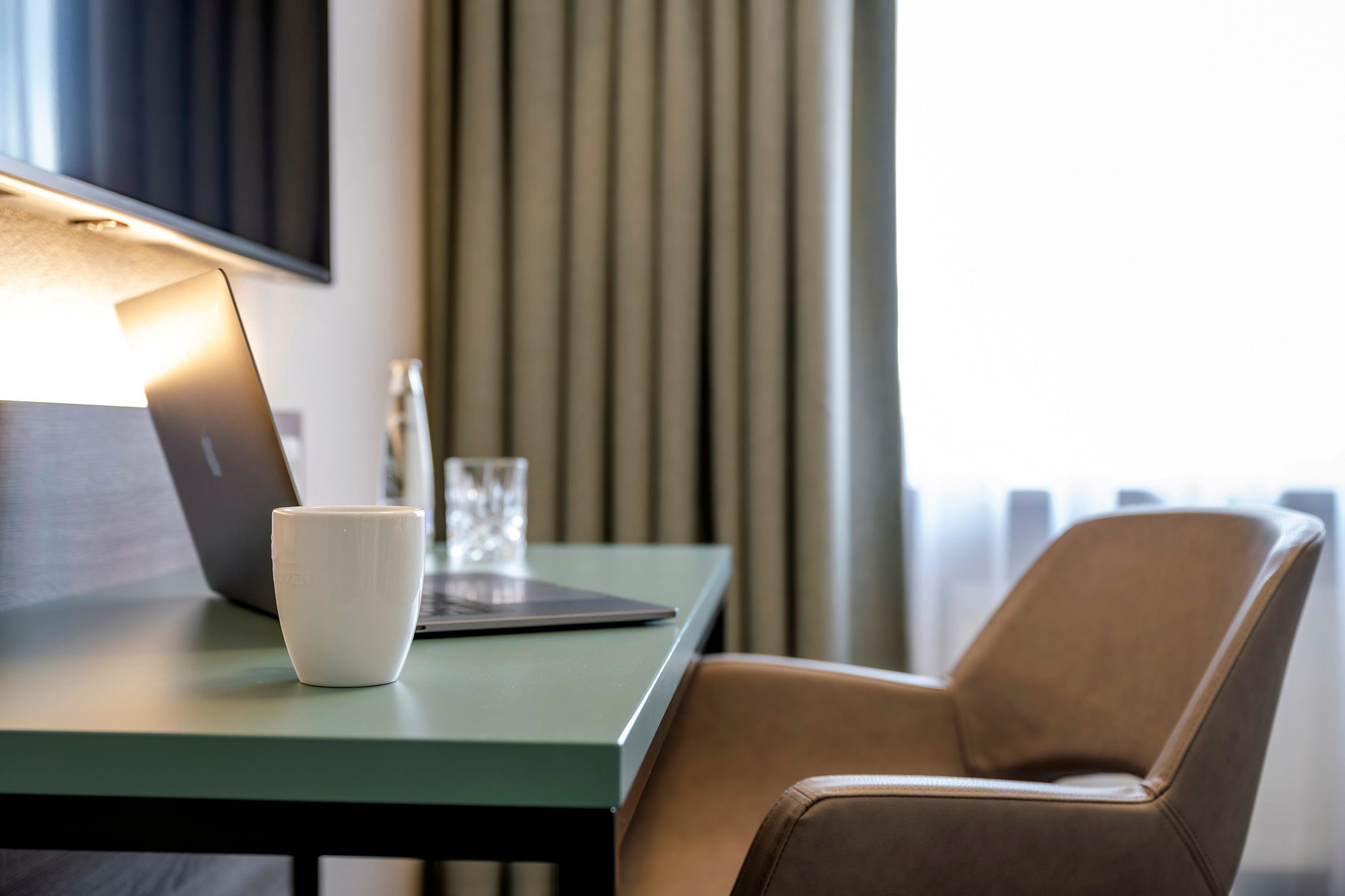 Una habitación moderna con una mesa verde, una computadora portátil, una taza blanca y un sillón de cuero marrón.